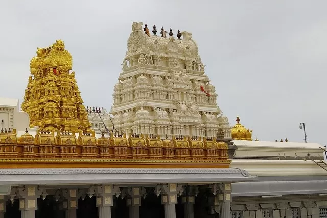 A daytime view of the roofs of Kamakshi Amman Temple in Kanchipuram, Tamil Nadu, showcasing the brilliant gold-plated vimana tower on the left alongside a multi-tiered white carved gopuram in the center. Golden decorative finials line the roof ledge above a pillared corridor against an overcast sky.