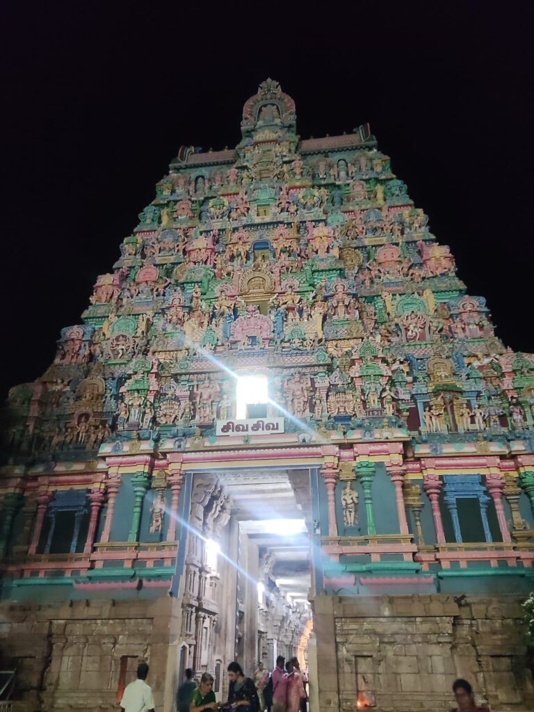 Colorfully illuminated gopuram and entrance corridor of Jambukeswarar Temple in Thiruvanaikaval, Tamil Nadu, with devotees entering at night.