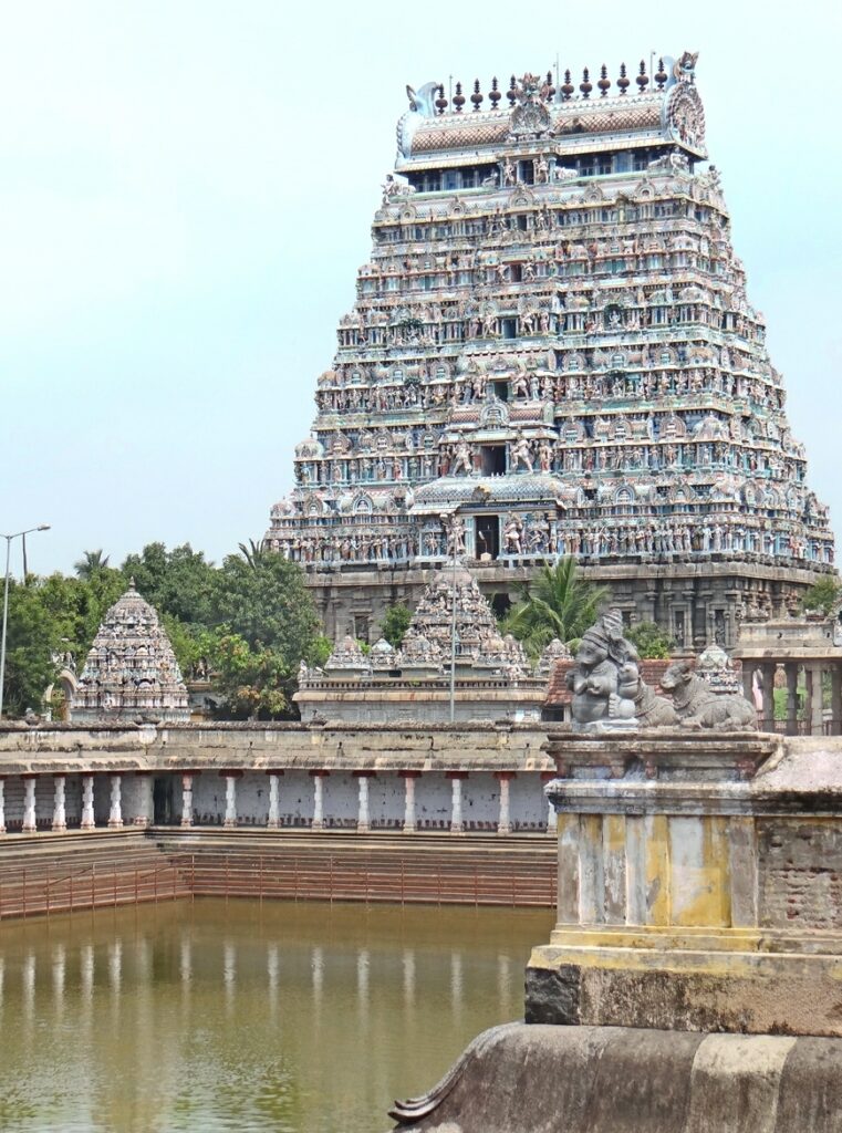 A high-angle view of the iconic multi-tiered, colorful gopuram (gateway tower) of the Thillai Nataraja Temple in Chidambaram, Tamil Nadu. In the foreground is the temple's sacred water tank (Shivaganga) surrounded by stone steps, with small stone carvings of Nandi and Ganesha atop a foreground pillar.