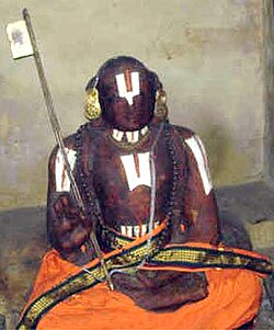 A seated idol of Sri Ramanuja at the Ramanuja Shrine in Ranganathaswamy Temple, Srirangam, wearing orange robes, tilak markings, and holding a tridandam staff.