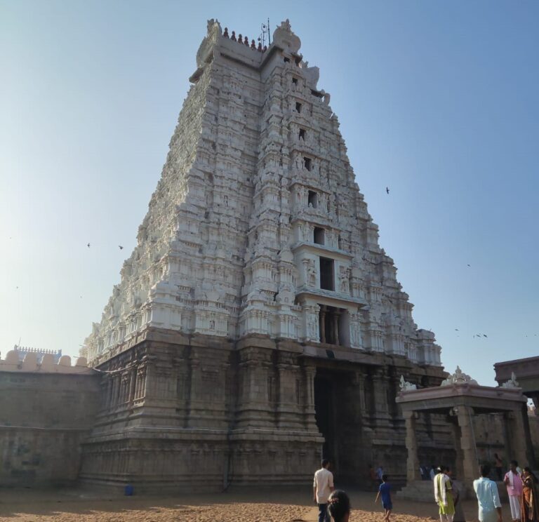 A low-angle shot showing the tall, intricately carved white gopuram (tower gate) of Sri Ranganathaswamy Temple in Srirangam, Tamil Nadu. The tower features detailed stone carvings and sculptures along its tiers, set against a bright, clear blue sky with birds flying around. Visitors are seen walking around the stone courtyard near the entrance at the base.