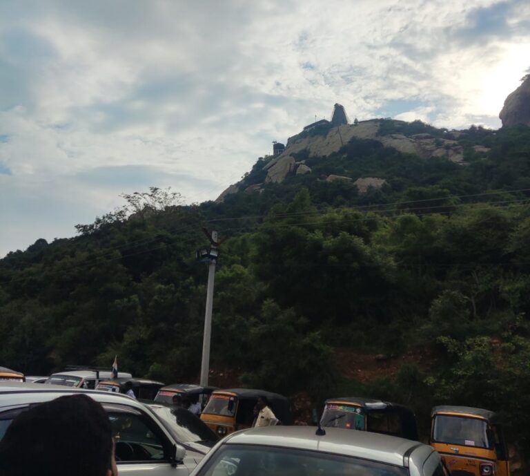 Rocky hilltop of Sholinghur temple in Tamil Nadu, viewed above a busy parking area with cars and auto-rickshaws.