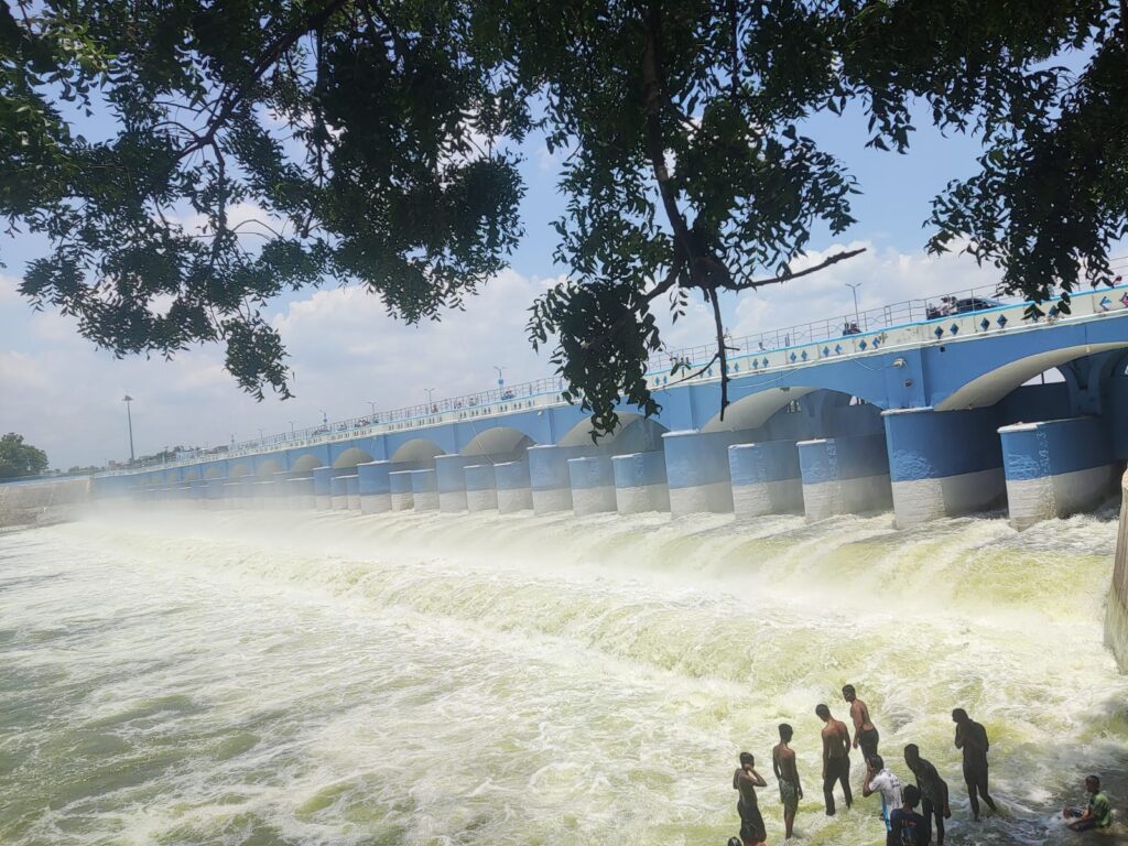 A wide view of the historic Kallanai Dam (Grand Anicut) in Tamil Nadu under a partly cloudy sky, framed by tree branches above. White foam and rushing water spill over the blue and white arched bridge structure. In the foreground, a group of young men stand in the shallow, fast-flowing water at the base of the dam.