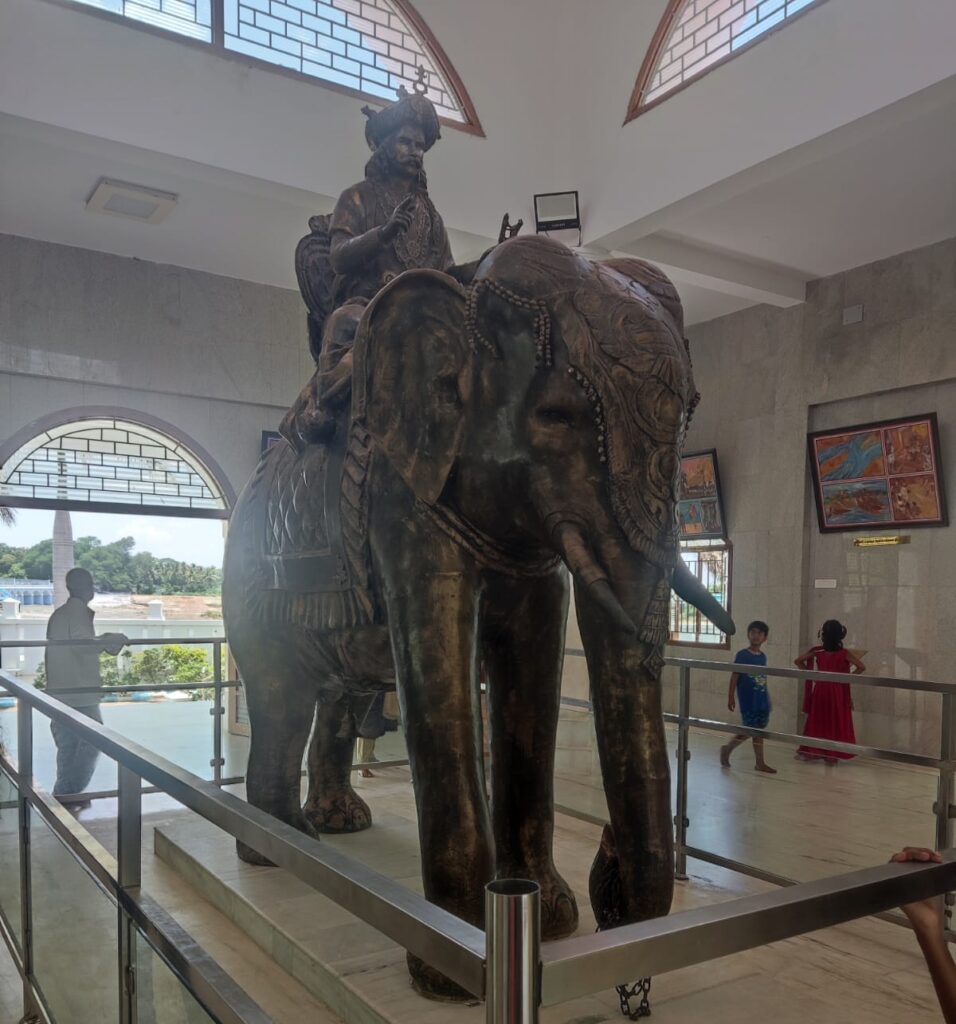 A large bronze statue of the Chola king Karikala Chola seated atop a decorated elephant, displayed inside the memorial hall at Kallanai Dam, Tamil Nadu. The statue is enclosed by metal safety railings, with visitors walking around the tiled indoor gallery space in the background.