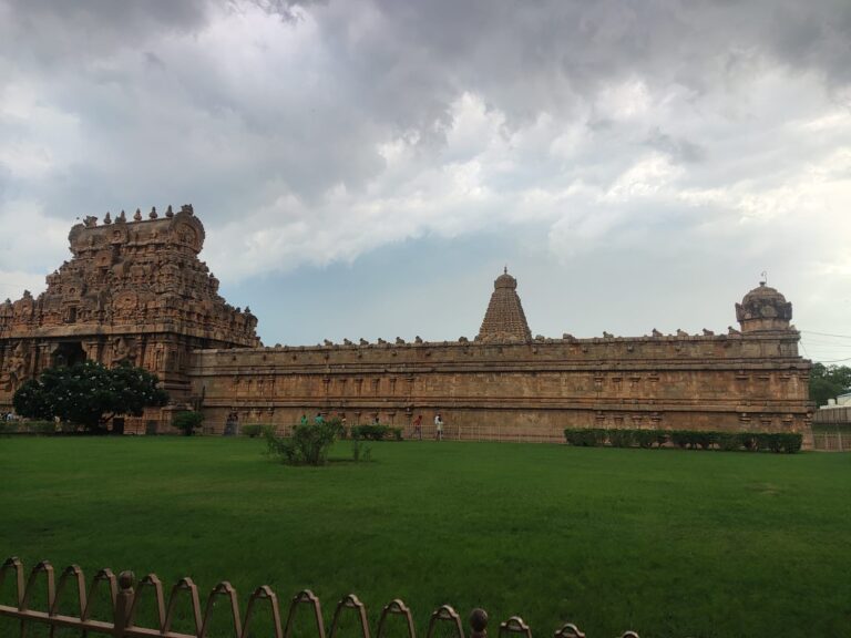 A wide side view showing the sprawling stone compound wall, entrance tower (gopuram), and main vimana apex of the Brihadeeswarar Temple in Thanjavur, Tamil Nadu. A neat green lawn fills the foreground, set beneath a dramatic, dark overcast sky.