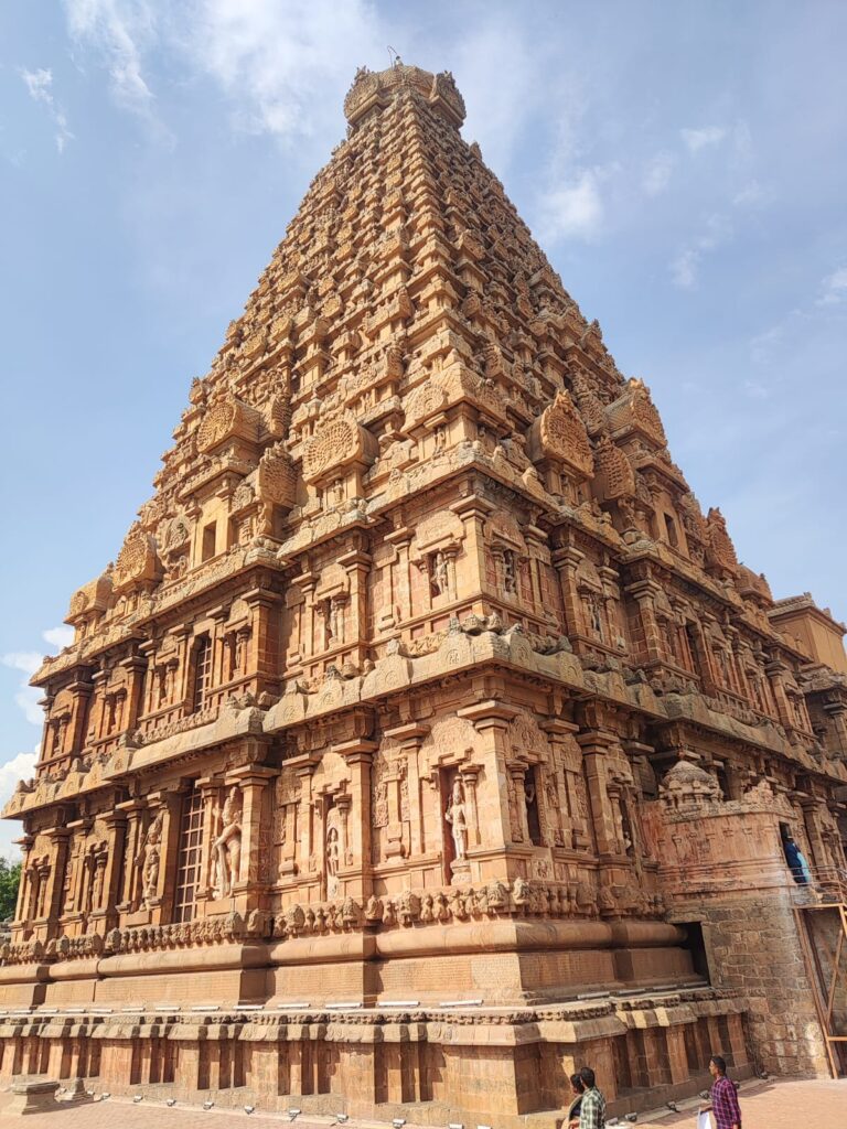 A low-angle, detailed view of the massive pyramidal vimana (main tower) of Brihadeeswarar Temple, showcasing intricate Chola-style stone carvings, pillars, and deity relief sculptures stacked along its multi-tiered brown granite facade under a clear blue sky. we found one of most beautiful architecture at brihadeeswarar temple in our tamil nadu temple tour