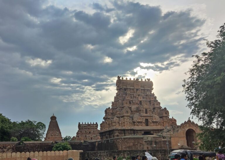 A shot of the entrance gopurams of Brihadeeswarar Temple in Thanjavur, with the tip of the central vimana visible in the background against a wide, cloud-filled sky.