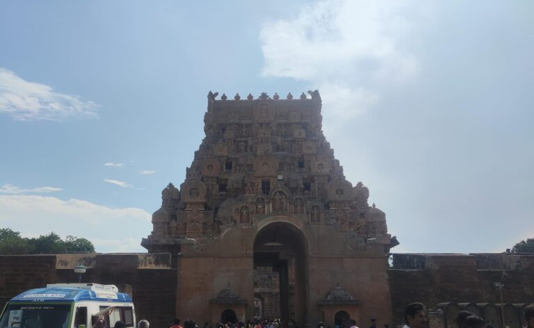 A direct front-facing view of the entrance gopuram at Brihadeeswarar Temple, displaying its large arched gateway and decorative stone tiers. Visitors and a mobile ATM van are visible near the outer complex entry in the foreground.