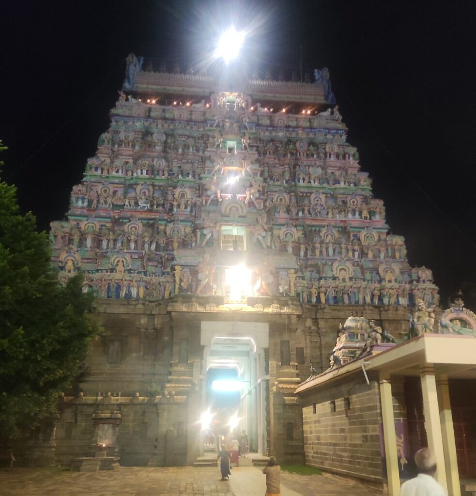 A nighttime shot of a brightly lit, multi-tiered gopuram (tower gate) at a temple in Kumbakonam, Tamil Nadu. The intricate sculptures and vibrant colors on the tower layers are illuminated by strong floodlights, with light glaring from the center. People are seen walking in the stone courtyard leading into the temple's entrance passage.