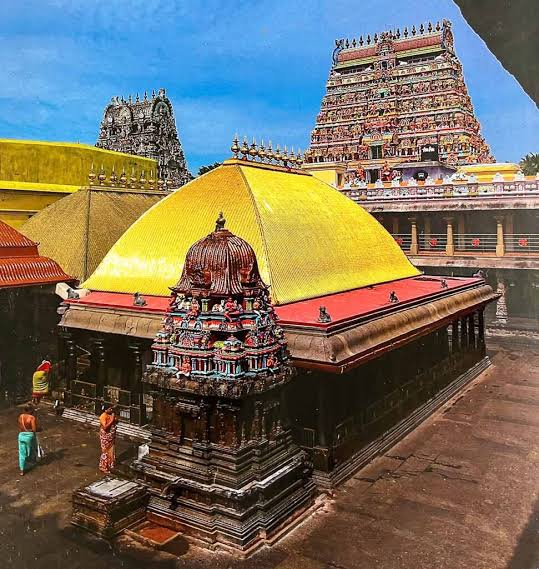 A vibrant view showing the signature gold-tiled roof (Kanaka Sabha) of the Chidambaram Nataraja Temple inner shrine. In the background, the tall, colorful gopuram stands against a bright blue sky, while devotees walk in the surrounding stone courtyard below.